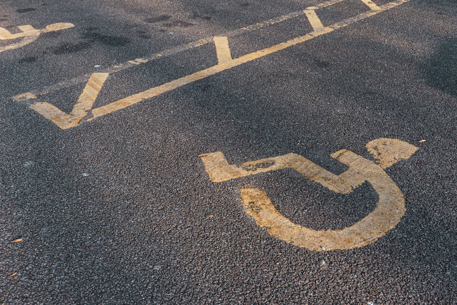 A close-up view of a textured asphalt surface showing a white-painted disabled parking space symbol, featuring a wheelchair icon and diagonal lines. The paint appears weathered with some chipping and fading, and the surrounding pavement is dark with small pebbles. Inside the parking space outline, the ground is clear of obstructions. This setting is outdoors, hinting at a parking area designated for accessible parking, likely near a property or commercial premises in East Dulwich. The image highlights the importance of clear signage for parking access, relevant to house removals and moving logistics, such as those managed by Movers Dulwich, during home relocation or furniture transport processes requiring convenient parking access.