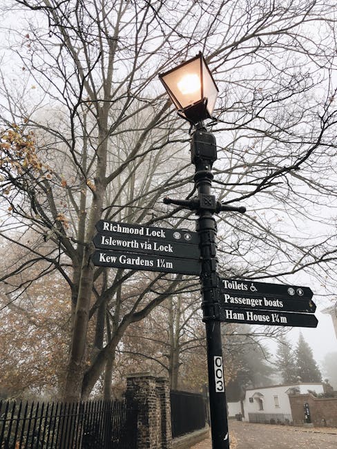 A tall black metal street lamp with a lit lantern at the top stands in front of leafless trees on a foggy day, with multiple directional signs attached to the post. The signs display destinations such as 'Richmond Lock,' 'Isleworth via Lock,' and 'Kew Gardens 1/4m,' indicating nearby locations and walking distances. In the background, there is a brick fence and a small white building with a pitched roof, partially obscured by the fog. The scene is typical of a residential or park area in Dulwich, with an overcast atmosphere. Movable boxes, packing blankets, or moving equipment are not visible in this image, but the setting suggests an environment where house removals and furniture transport may take place. The image is suitable for describing the logistical aspects of home relocation and local moving services in the SE21 Dulwich area, as provided by Movers Dulwich, with a focus on outdoor signage and street scene context.