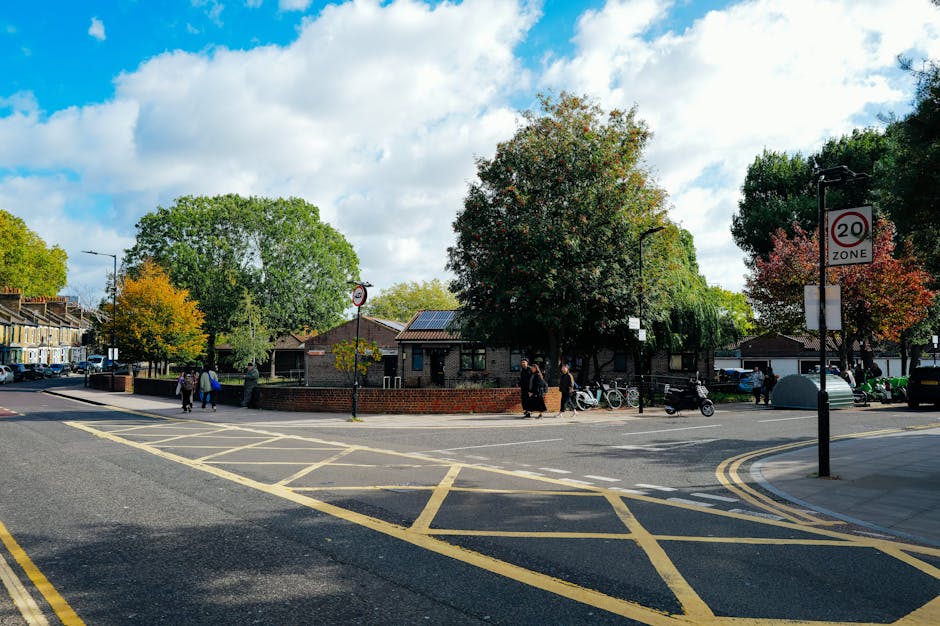 A view of a residential street in Dulwich with a central open area surrounded by trees showing autumn foliage. The scene includes a brick wall, a small building with solar panels on the roof, and several people walking along the pavement. Some pedestrians are carrying shopping bags or pushing bicycles, while others stand near parked bikes and scooters. The street features a crossing with yellow lines and a '20 zone' traffic sign, with a parked car and additional vehicles further down the road. Bright daylight and partly cloudy skies illuminate the scene, providing clarity for understanding the typical environment encountered during home relocation or furniture transport activities, as handled by Movers Dulwich in their removals and moving services.