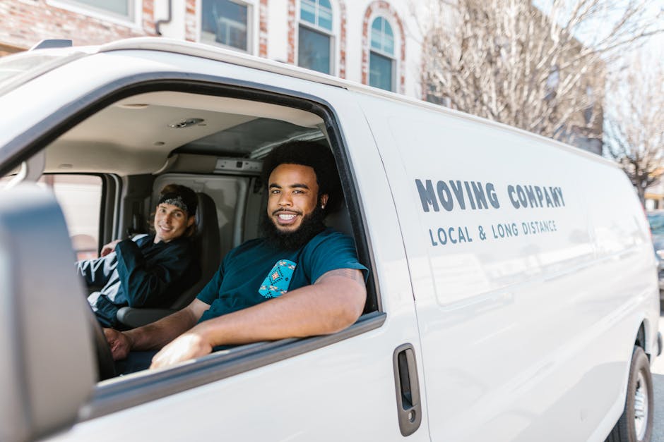 Two movers from Movers Dulwich are engaged in a home relocation process, loading a large piece of furniture wrapped in green moving blankets into the open back of a white van parked on a street near N