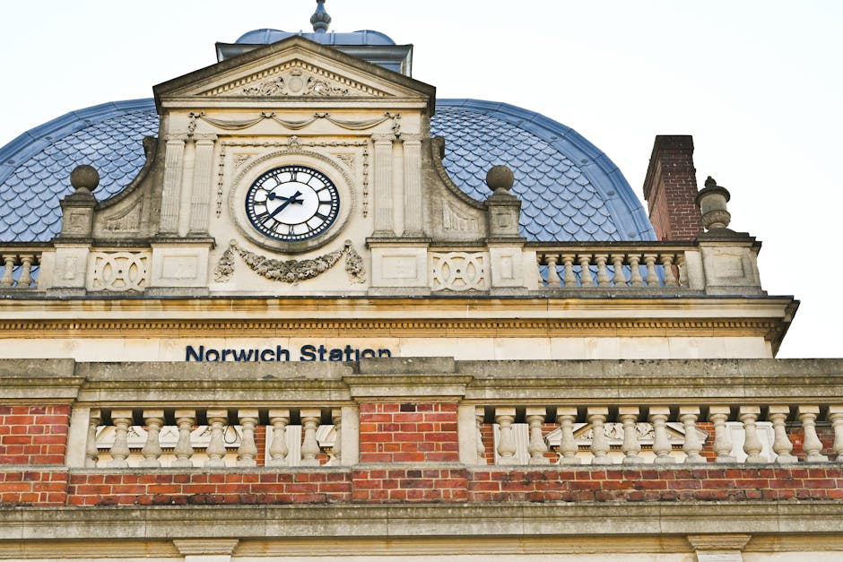A close-up view of the ornate upper facade of Norwich Station, showcasing its historic architectural features including a large round clock with black Roman numerals and hands, set in an elaborate stone frame. The building's exterior combines stone and brick elements, with a decorative stone balustrade running along the edge of the roofline, supported by classical stone columns and finials. Inside the station, visible through the image's perspective, there are no furniture or moving boxes present; it focuses on the station's historic design elements and the roof's blue-grey tiled dome. This image, associated with house removals and relocation services, emphasizes the careful handling of packing and transport processes, such as loading or unloading items, within a building of this type. Occasionally, Movers Dulwich may utilize such imagery to highlight the importance of professional furniture transport and home relocation logistics around historic station structures, like Norwich Station, in their services.