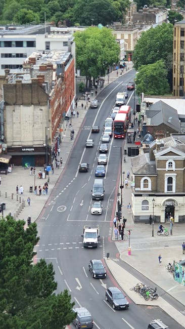 An aerial view of a city street during daytime showing a line of vehicles, including vans, cars, and a red bus, traveling along a curved road. The scene includes pedestrians walking on sidewalks, some carrying boxes or bags, and cyclists near a small building with a pitched roof. On the left side, a multi-storey building with large windows and a brick façade is visible, along with other older and modern structures. Trees with lush green foliage line the street, providing shade. In the foreground, a white van is approaching an intersection, and in the background, the street continues past more buildings and mature trees. The environment suggests a bustling urban area suitable for house removals and furniture transport, with evident activity of loading or unloading in proximity to residential and commercial locations, highlighting logistics involved in home relocation services provided by Movers Dulwich.