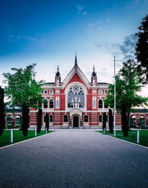 A large, ornate red brick building with decorative white trim, featuring a central arched window with intricate stained glass and multiple smaller windows. The structure has a steeply pitched roof wit